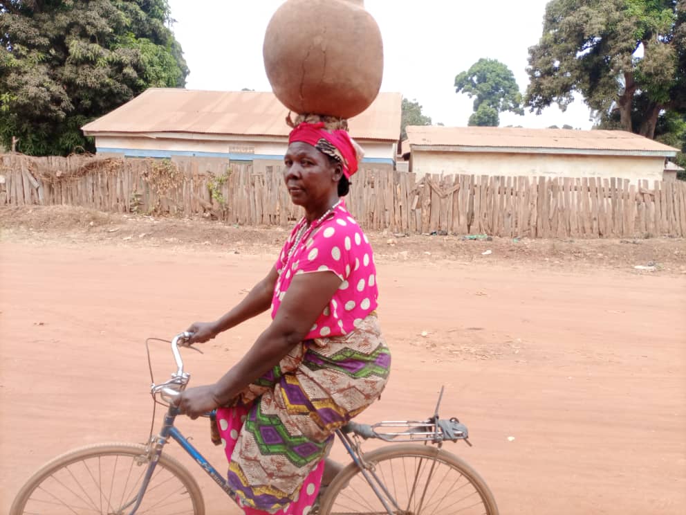 Yambio woman rides bicycle with pot on her head